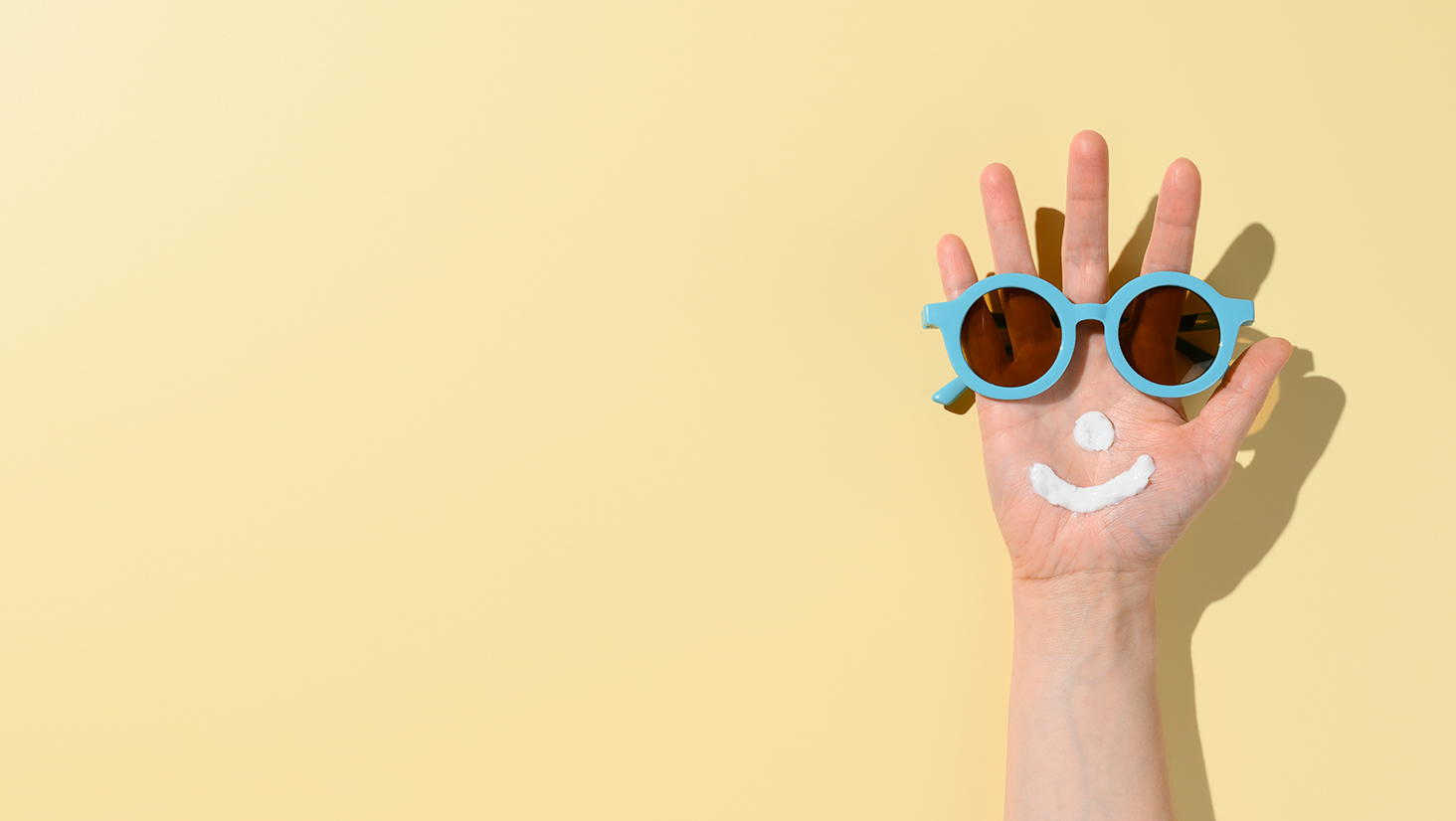 Funny smiling face created from sunscreen cream and sunglasses on female hand. Minimal summer scene on sandy-colored background. Health care and beauty concept. Copy space, banner
